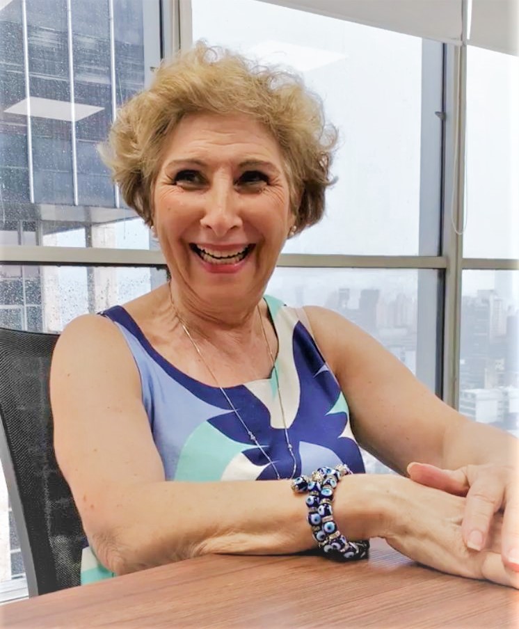 Profissional veterana sentada em uma cadeira com as mãos na mesa, sorrindo. Ao fundo, uma janela na qual há uma vista da cidade de São Paulo