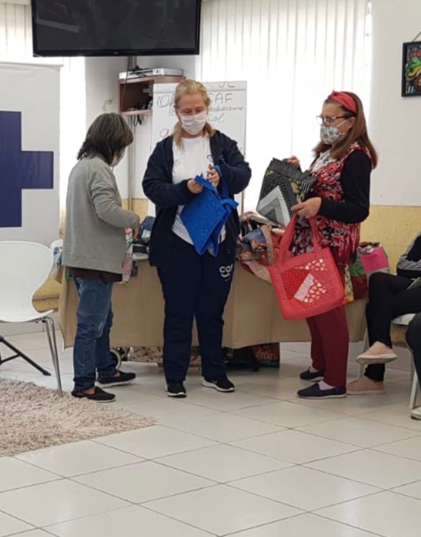 Três mulheres presentes na foto, a primeira esta de lado olhando para a mesa que possui artesanatos em cima, a do meio esta segurando uma bolsa azul, e Dona Maria está segurando duas bolsas uma vermelha e uma cinza. 