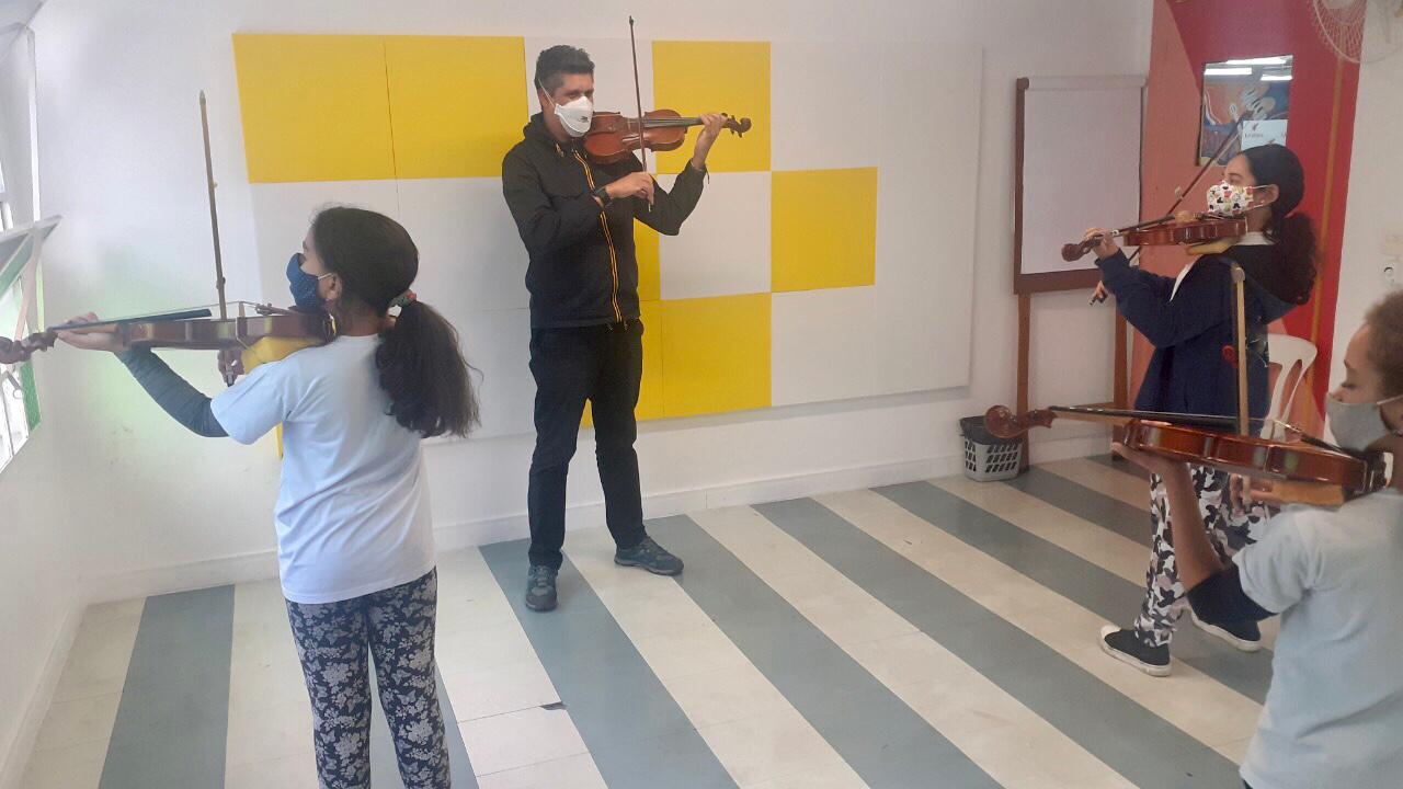 Uma sala com chão listrado e uma parede com quadrados amarelos. Em frente a essa parede tem o professor de máscara segurando o seu violino. Diante do professor tem três crianças de máscara e cada uma delas segura o próprio violino. 