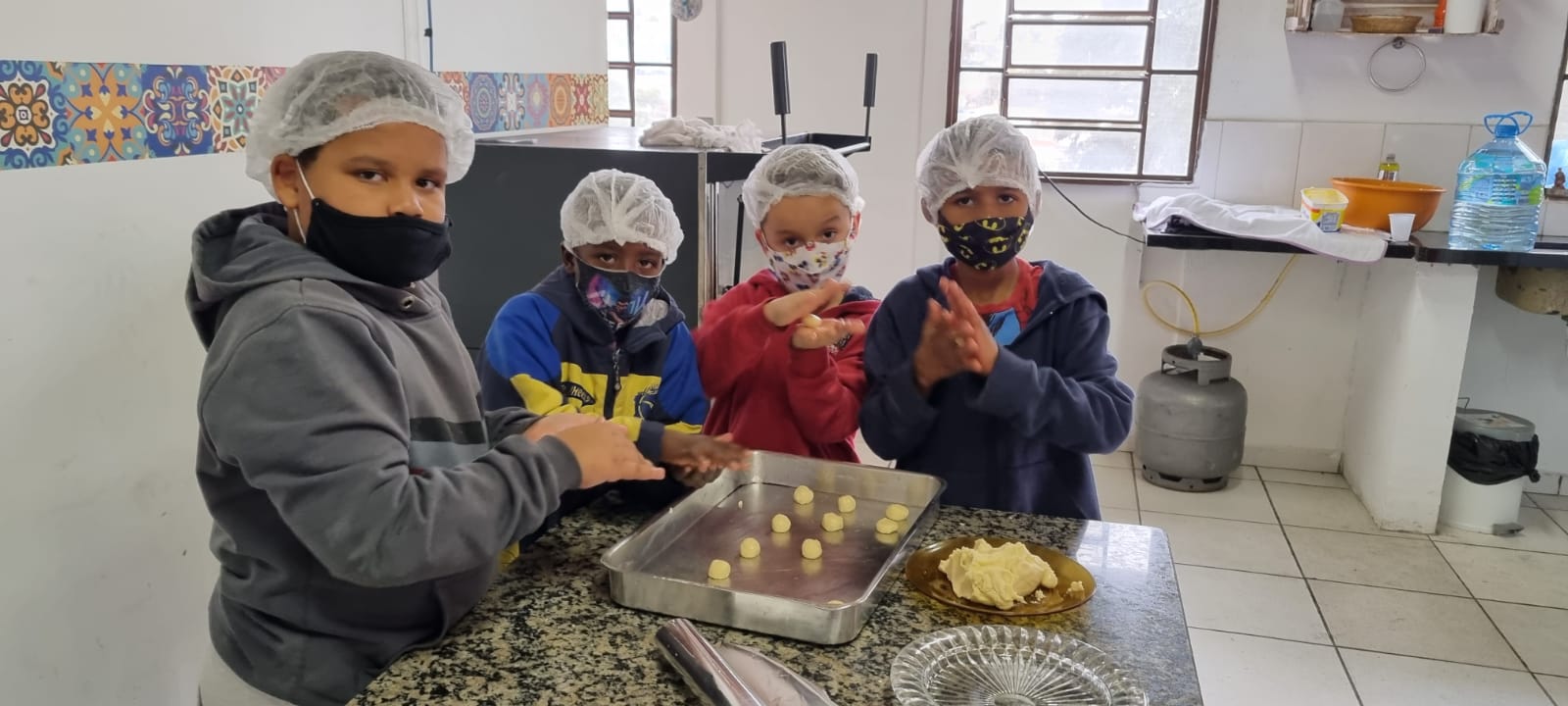 Quatro crianças de máscara e touca higiênica em uma cozinha preparando biscoitos de maizena. Na frente deles tem uma mesa com uma forma e pratos com a massa do biscoito.  