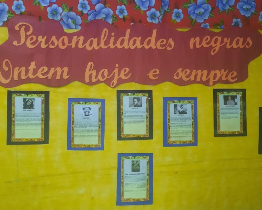 Mural vermelho e amarelo com seis folhas distribuídas com o nome de figuras públicas negras. Na parte de cima em vermelho está escrito “Personalidades negras ontem, hoje e sempre”