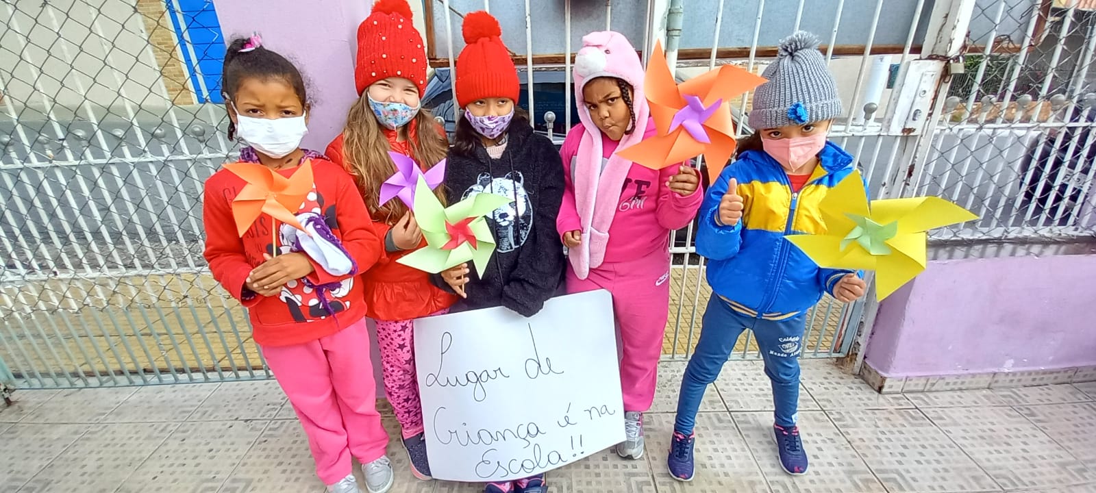 Cinco crianças reunidas, cada uma segurando um cata-vento, símbolo da campanha, a criança do meio está segurando um cartaz escrito “lugar de criança é na escola”.