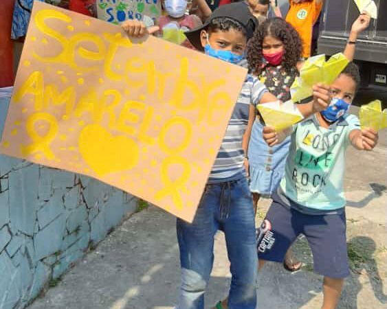 Uma criança de máscara segurando um cartaz escrito “Setembro Amarelo” ao lado e atrás dele tem crianças de máscara segurando um coração amarelo.  