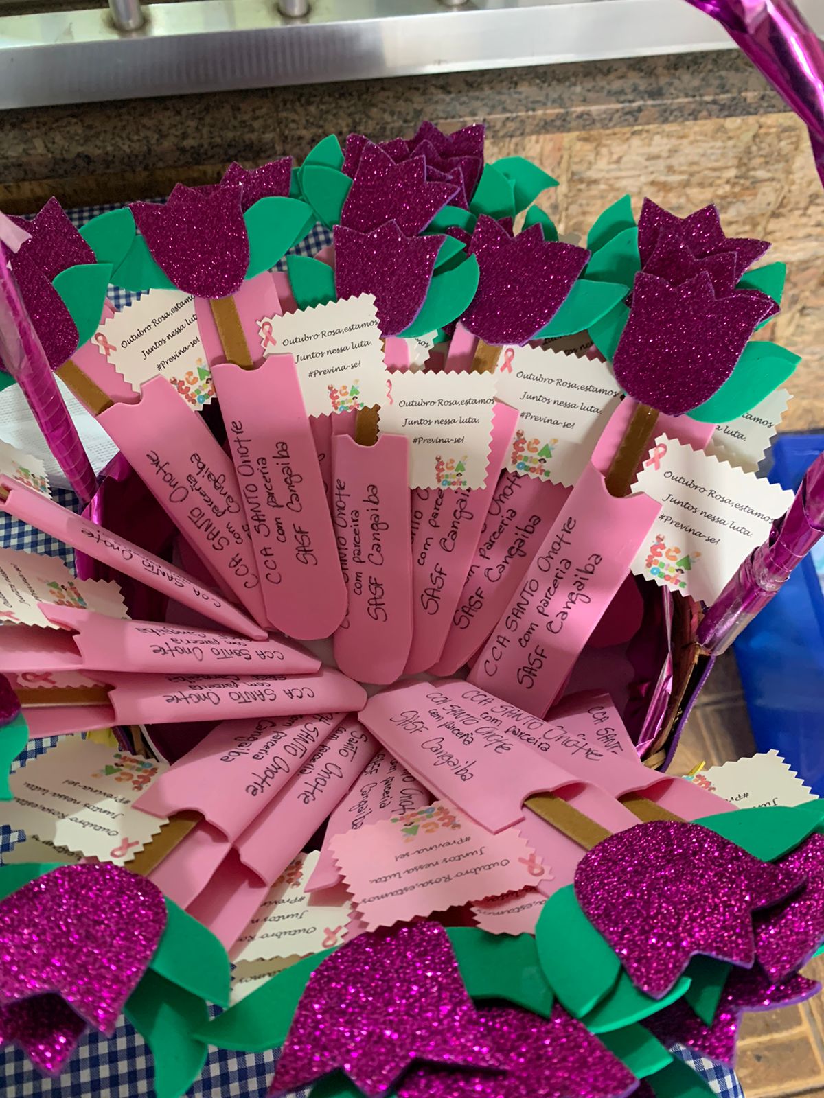 18 flores de pétalas E.V.A e cabo de palito de sorvete, com um bilhete colado em cada flor sobre a prevenção ao câncer de mama, tendo menção aos dois serviços organizadores do evento escrito em preto no cabo da flor. 