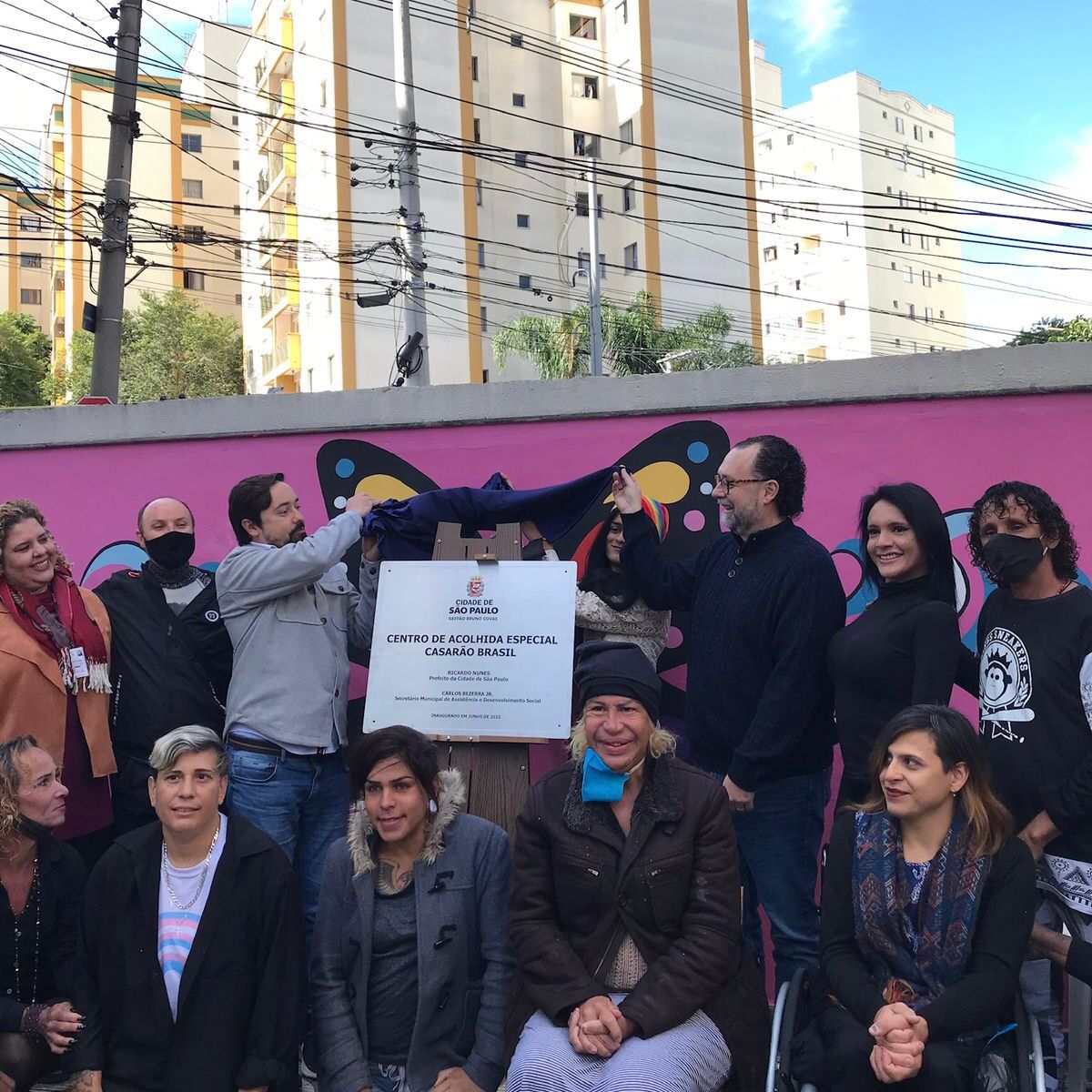 12 pessoas em volta da placa de inauguração do Centro de Acolhida Especial (CA) para mulheres trans - “Casarão Brasil”