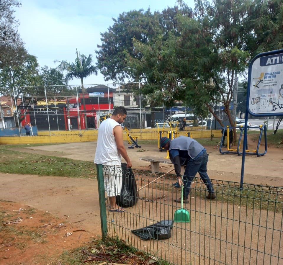 Um homem de colete junta a terra de um dos canteiros da praça enquanto outro homem aguarda segurando um saco de lixo preto