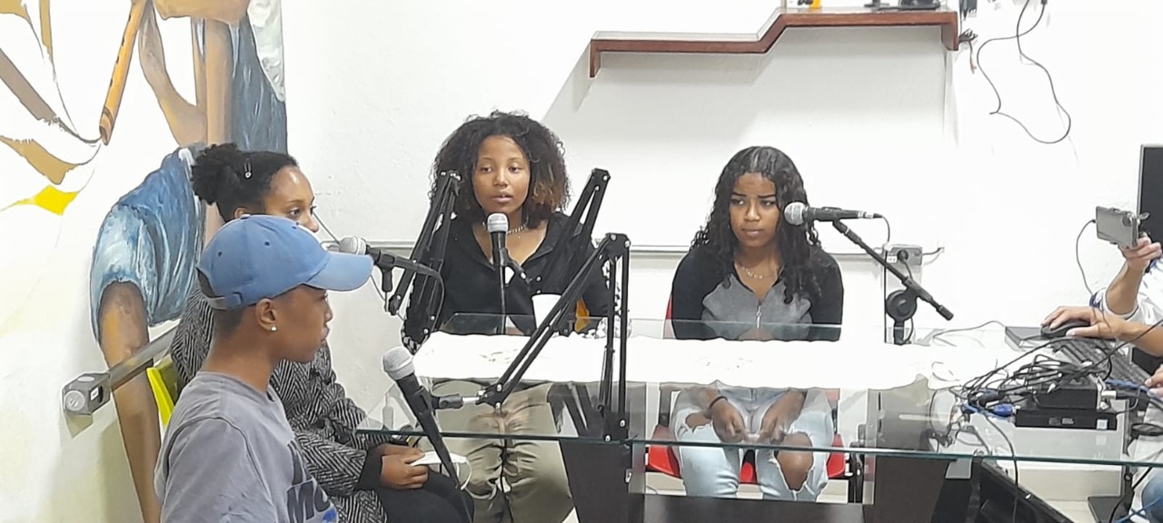 Quatro jovens sentam em cadeiras, de frente para uma mesa de vidro com microfones na superfície. No canto direito, uma mão segura o celular que grava o podcast.