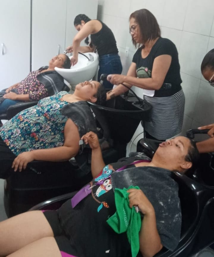 Na imagem, algumas mulheres estão lavando os cabelos de meninas do NAISPcD e de mães que estão sentadas em cadeiras-lavatório.