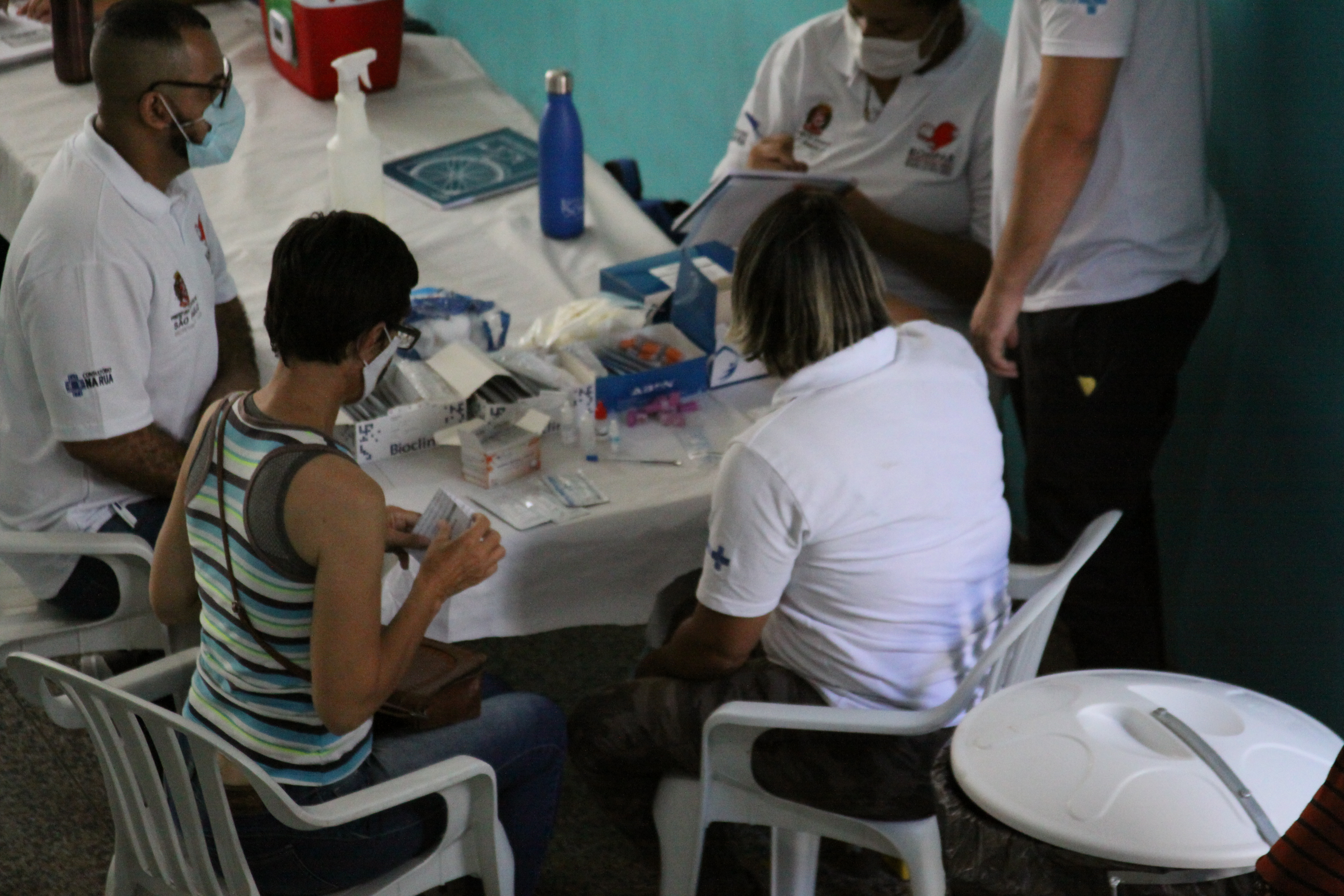 Dois agentes de saúde e uma paciente sentados em cadeiras em frente a mesa com material de coleta de exames e um agente de saúde em pé