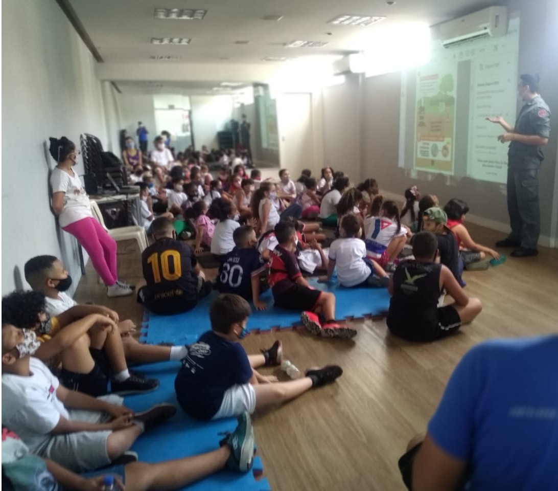 Dezenas de crianças sentadas em tapetes azuis prestando atenção no palestrante que esta em pé de frente para todas as crianças.