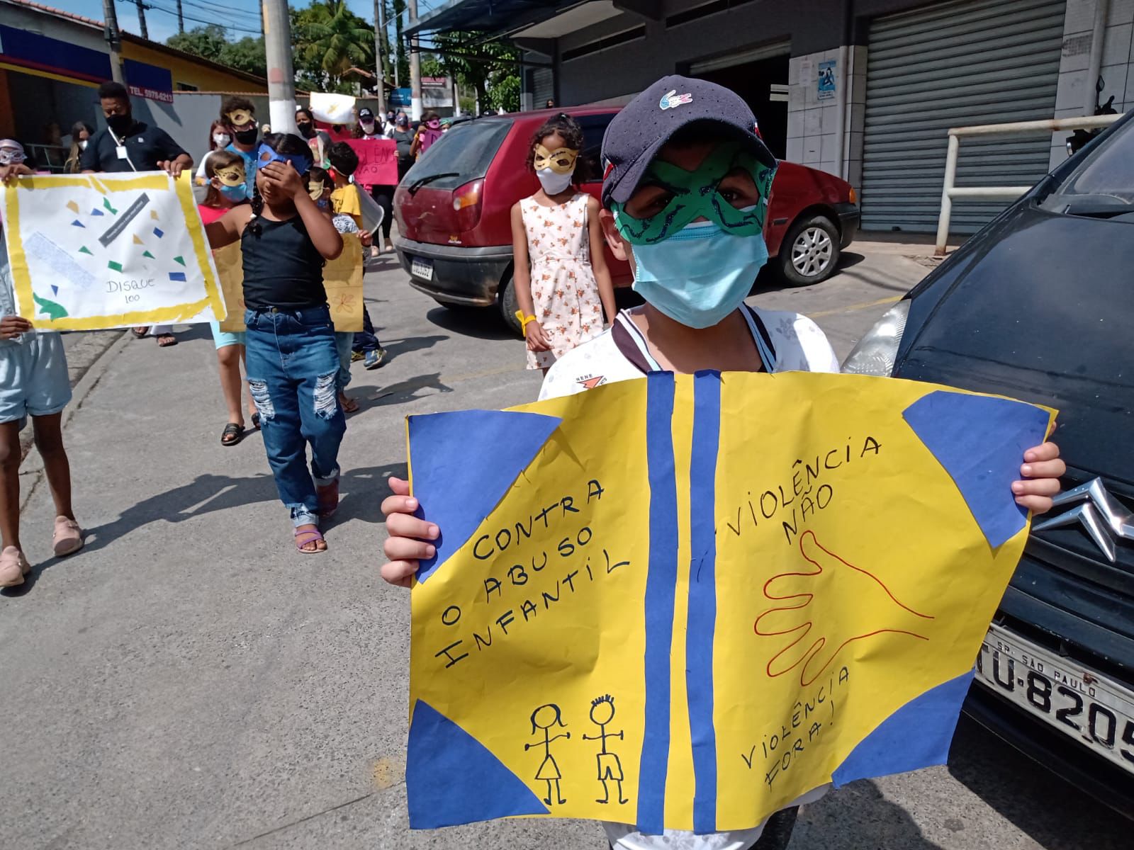 Em uma calçada com dois carros estacionados, crianças com máscaras de carnaval seguram cartazes contra o abuso e a exploração sexual infantil.
