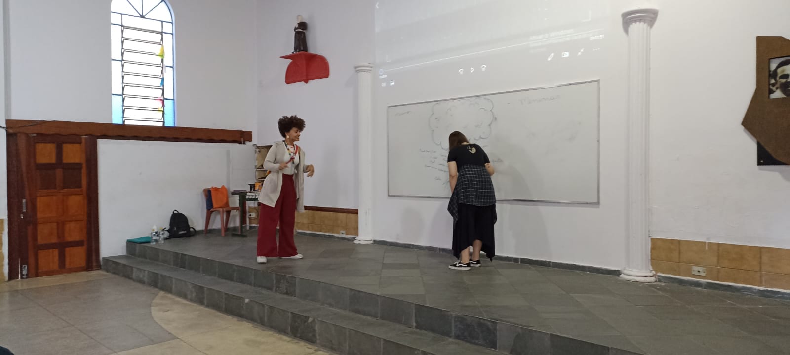 Duas mulheres estão em cima de um pequeno palco preto. Atrás delas há duas colunas brancas na parede. Na parede, há um quadro branco, onde elas fazem alguns desenhos e escrevem algumas palavras.