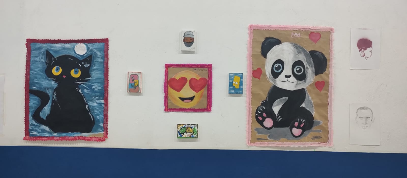 Na imagem, quadros são expostos em uma parede branca. O da esquerda, de um gato preto. Na direita, o desenho de um panda.