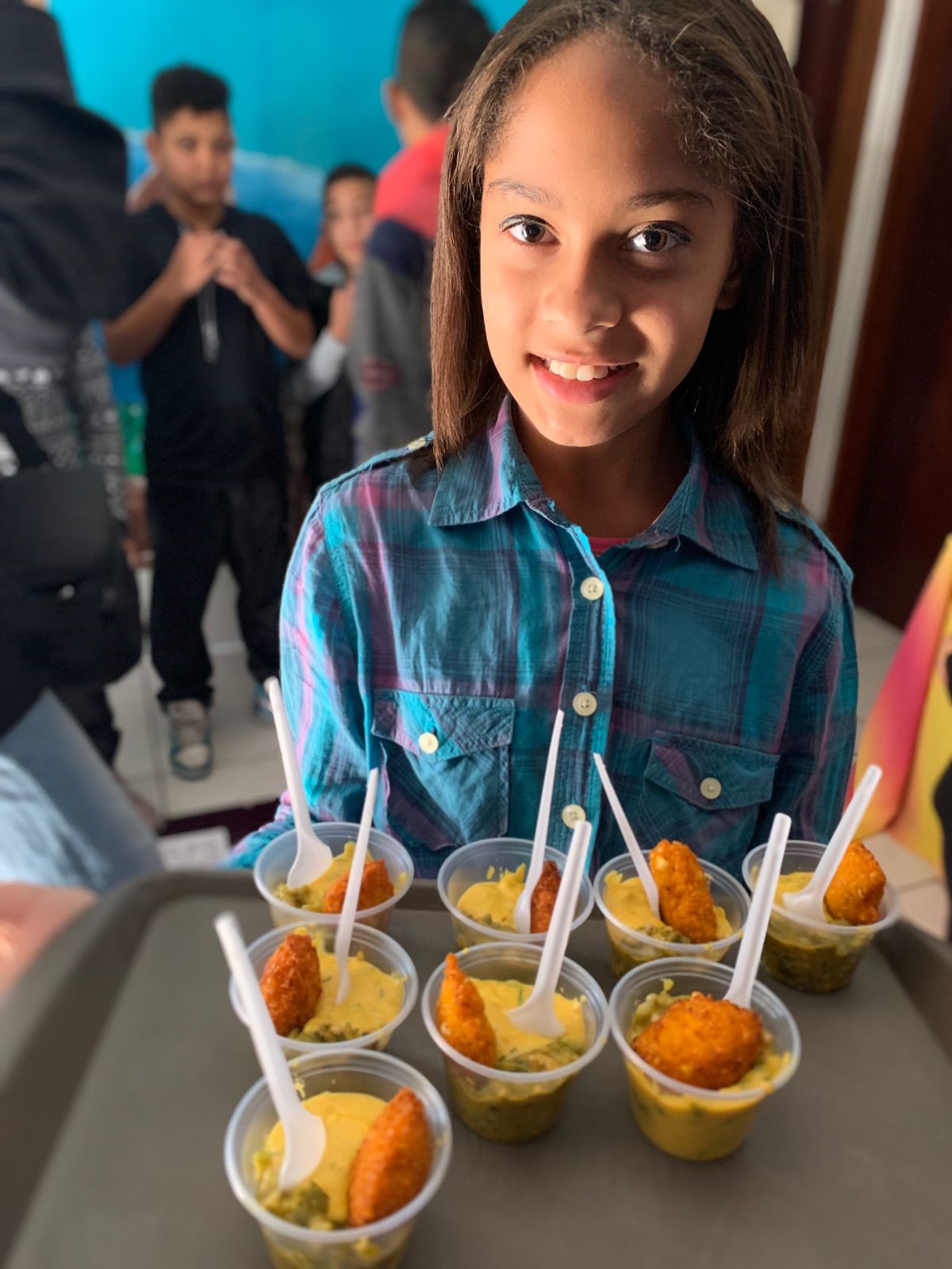 Criança do CCA Nazareth vestida com camisa xadrez representando o nordeste do país, segurando em suas mãos uma bandeja com mini acarajés, prato típico da Bahia. 