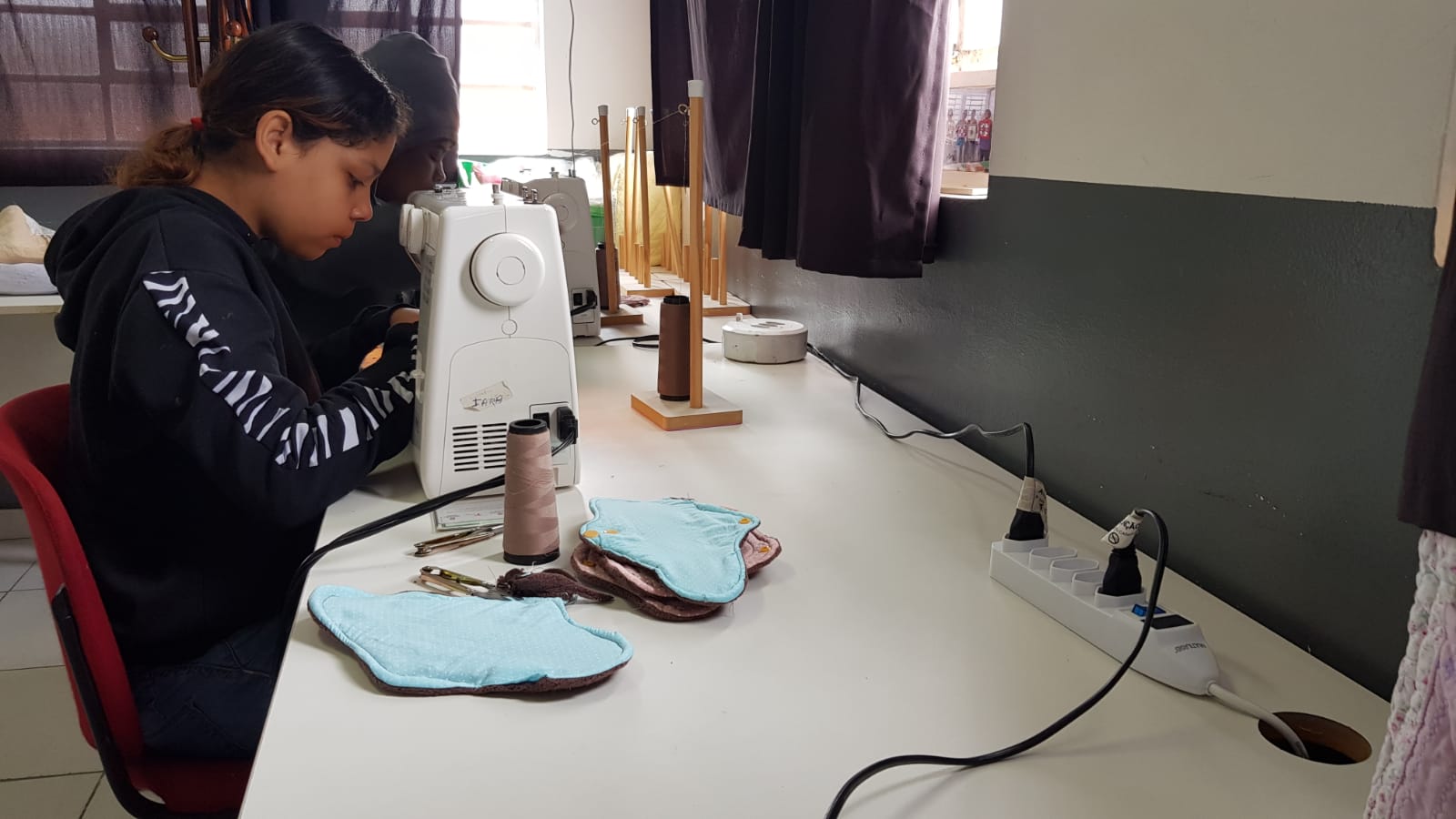 Duas meninas trabalham nos absorventes em máquinas de costura. Em cima da mesa já há quatro absorventes prontos, além de materiais de costura.
