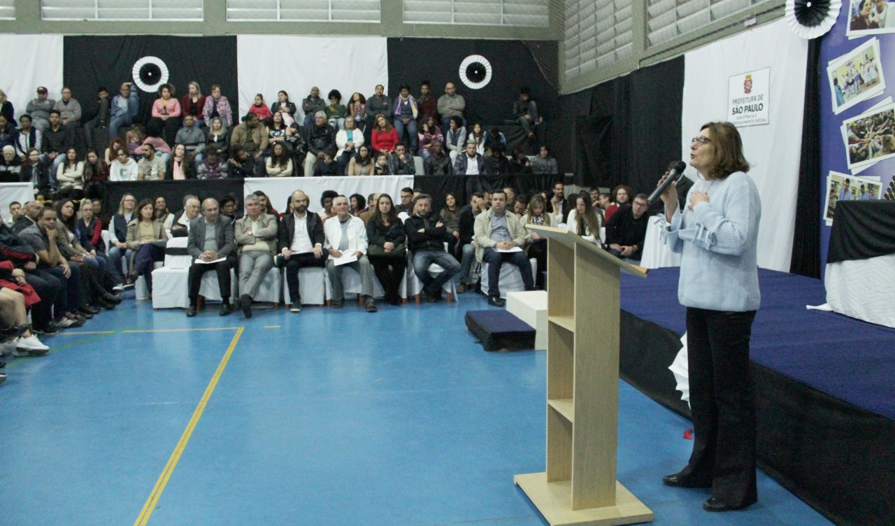 Secretária Berenice Gianella discursa com microfone na mão diante de plateia a sua frente e a sua direita em galpão do serviço. Atrás dela há um palco com fotos coladas em pano azul e o brasão da Prefeitura de São Paulo