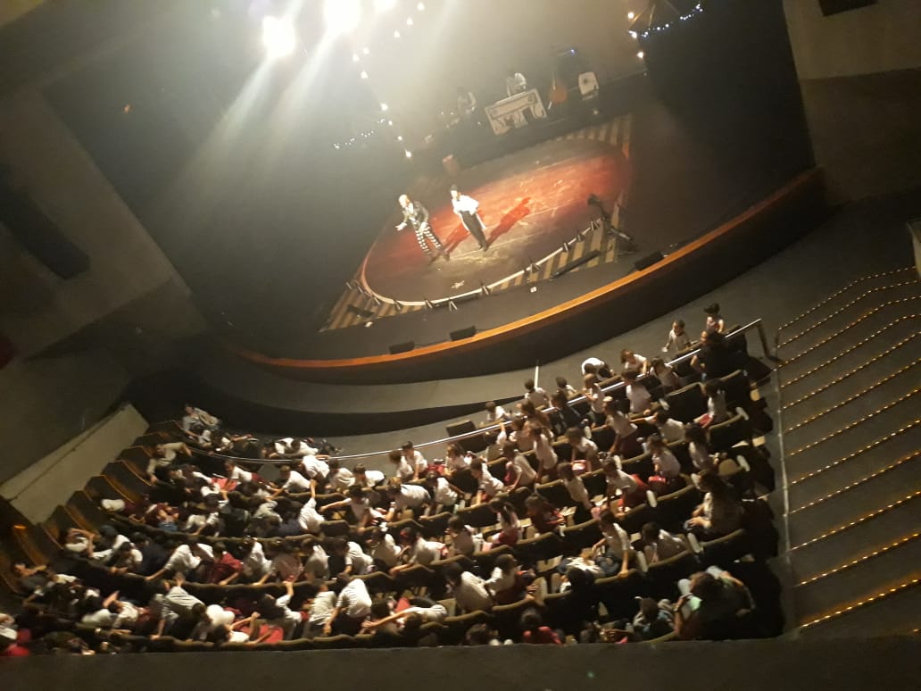 Plateia do teatro vista de cima com crianças sentadas nas poltronas e dois atores no palco