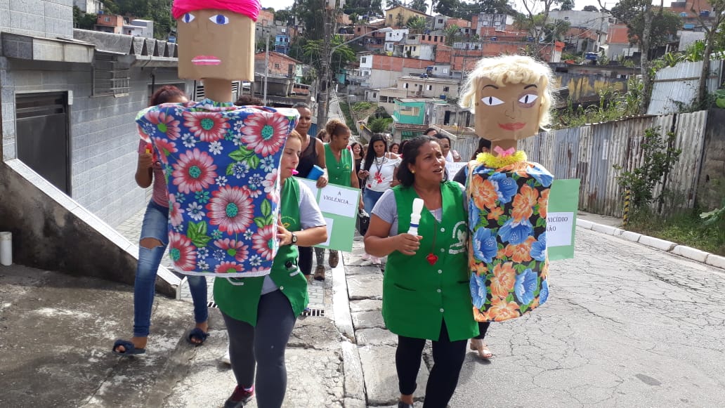 Duas funcionárias dos serviços, com coletes verdes, caminham à frente em marcha contra o feminicídio, segurando cada uma um bonecas grandes artesanais de papelão