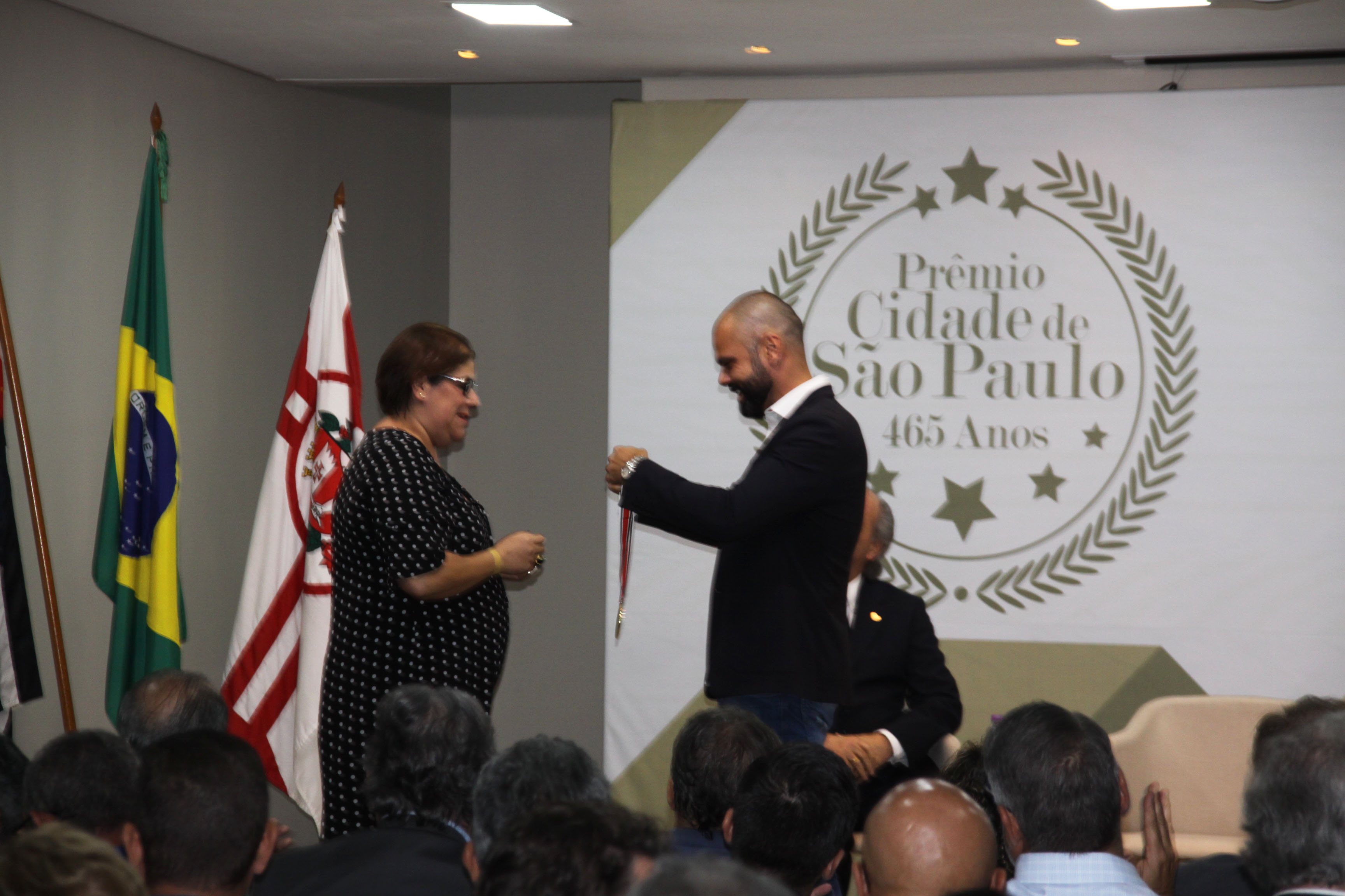 Coordenadora de Gestão SUAS no palco diante do prefeito Bruno Covas para receber de suas mãos a medalha do prêmio