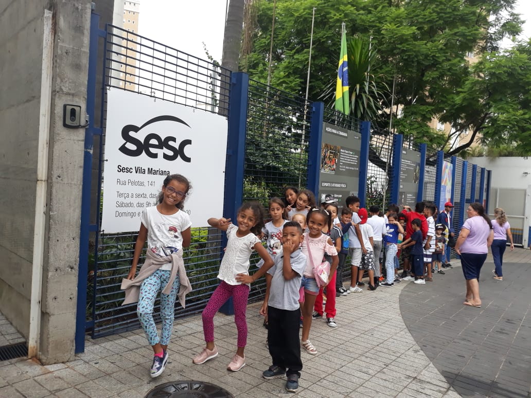 Crianças em fila na lateral externa do SESC Vila Mariana, com placa do local visível e três crianças a frente sorrindo na fila em pose para foto