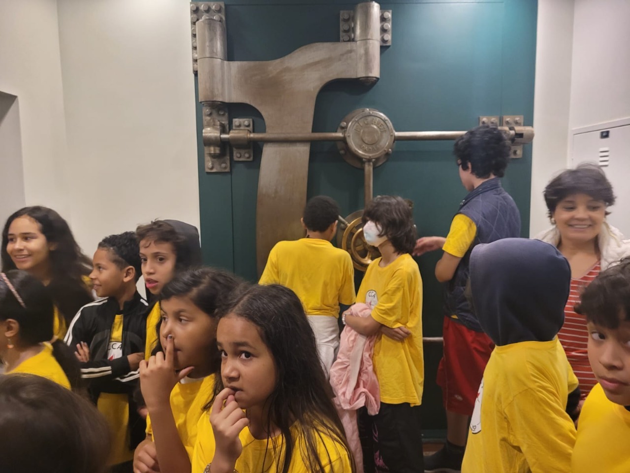 Crianças vestindo camisas amarelas estão na frente da porta de um Cofre. Ao fundo, duas crianças também de camisetas amarelas interagem com a porta do cofre.
