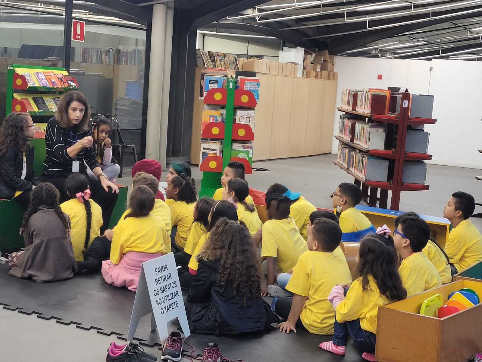 No brinquedoteca do Centro Cultural São Paulo, vestindo amarelo, crianças estão sentadas no chão enquanto a funcionária do Centro Cultural está contando uma história.