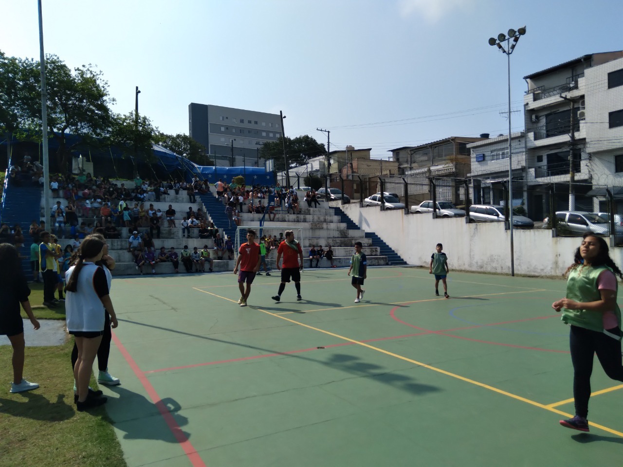 Em um dia ensolarado, crianças e adolescentes jogam futsal na quadra do Circo Social da Vila Ré, ao fundo, na arquibancada, os familiares torcem pelos jogadores.