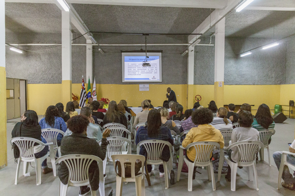 Dentro de uma sala, dezenas de pessoas estão sentadas em cadeiras assistindo à um seminário ministrado por um homem que está de pé e usando um microfone para falar.