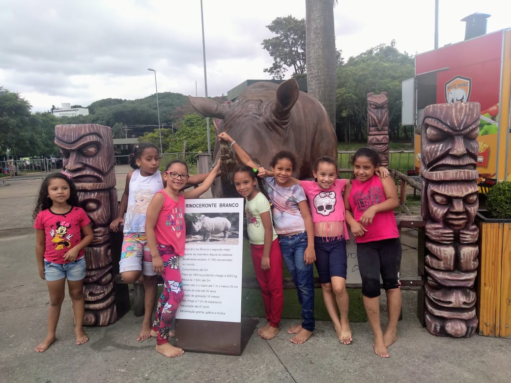 Sete garotas sorriem e posam para a foto. Elas estão de lado a lado na frente de uma estátua de um rinoceronte.