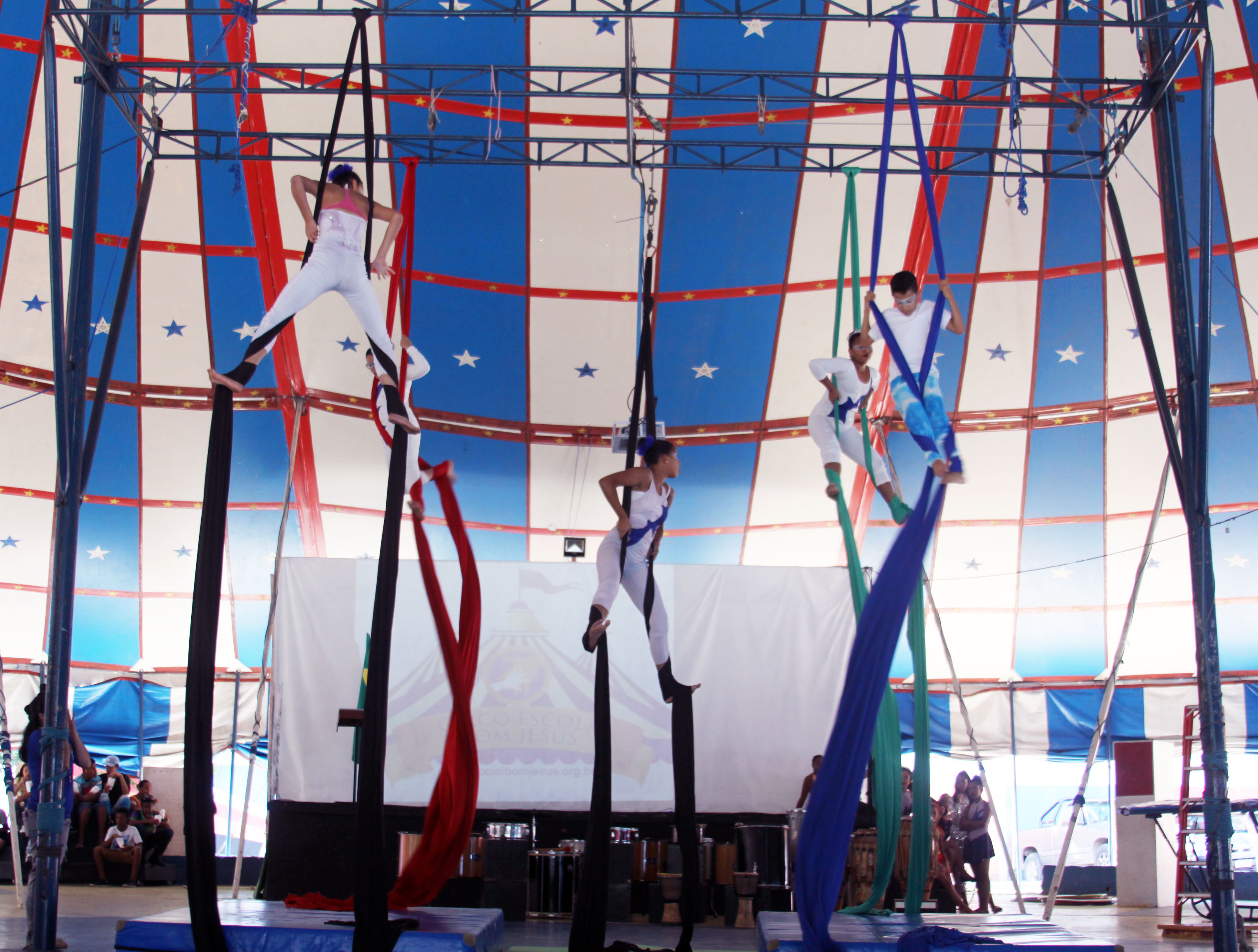 Dentro de uma lona de circo nas cores branca, vermelha e azul, cinco crianças se apresentaram com movimentos circenses enrolados em um tecido pendurado no teto.