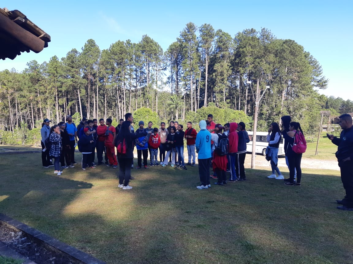 Cerca de 35 jovens em pé formando um semicírculo escutam instruções de um homem e uma mulher também em pé. Todos estão agasalhados ao ar livre próximos de muitas árvores.