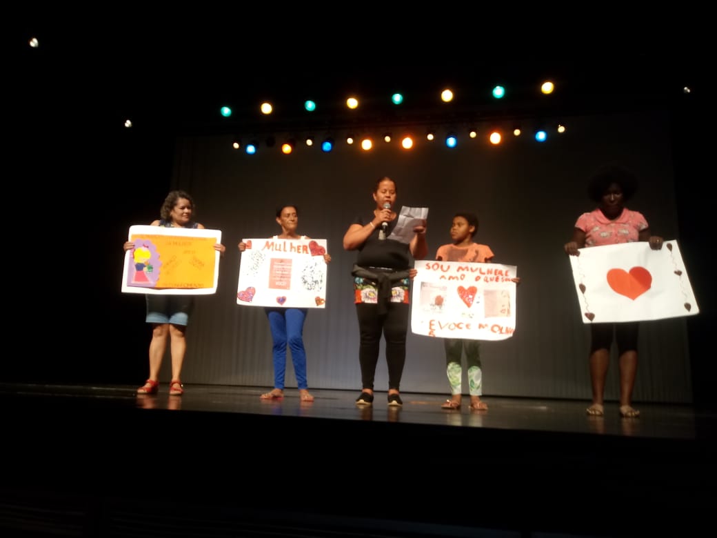 Em um palco, uma mulher de roupa preta está lendo homenagem às mulheres. Ao seu lado esquerdo estão 2 mulheres segurando cartazes decorados. Ao lado direito estão outras duas mulheres que também seguram cartazes decorados com coração. 