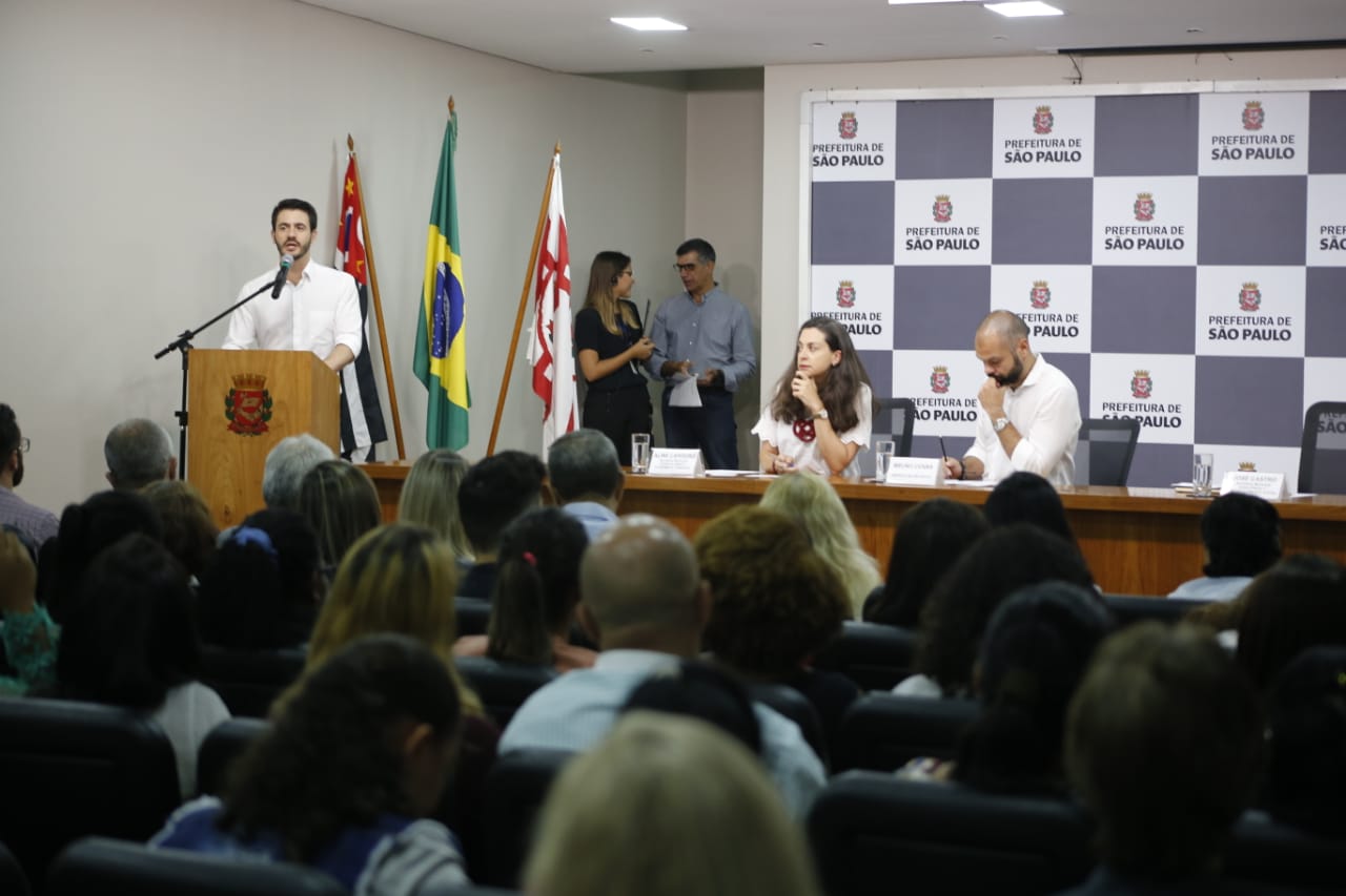 Secretário da SMADS, Jose Castro, fala ao microfone em auditório no canto esquerdo enquanto secretária da SMDE, Aline Cardoso, e Prefeito de São Paulo, Bruno Covas, estão sentados à mesa. 