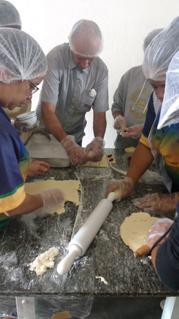 Em uma mesa de mármore, conviventes e oficineiro estão com touca e luva de higiene enquanto moldam a massa do biscoito e colocam o recheio 