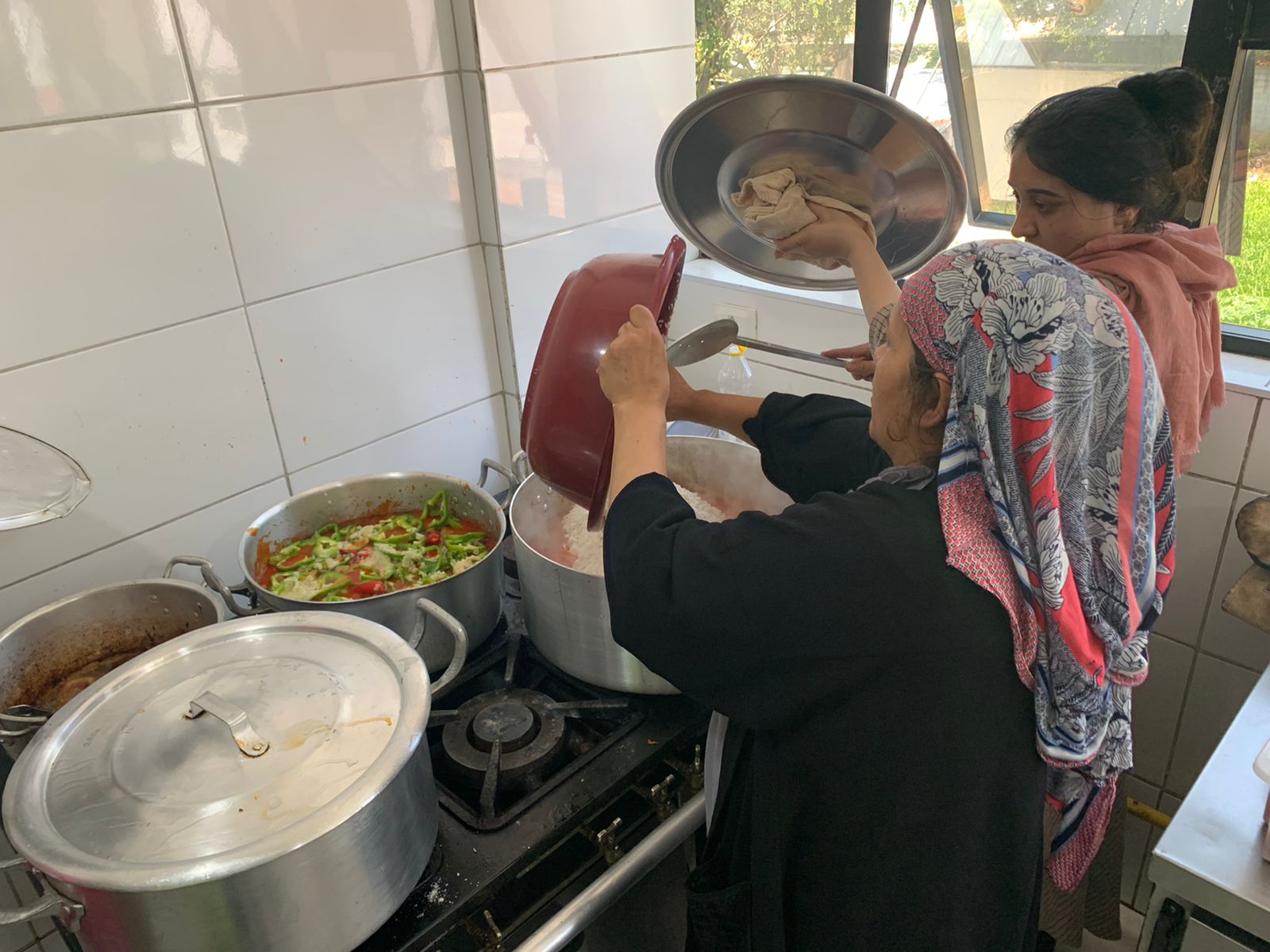 Duas mulheres afegãs estão rente ao fogão preparando a comida. Estão misturando diferentes ingredientes em uma grande panela. Usam lenços, para cobrir seus cabelos. 