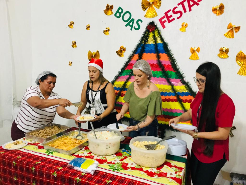 Quatro mulheres estão servindo seus pratos. A mesa tem várias opções de alimentos. Na parede ao fundo, há decorações de natal e frase “Boas Festas” escrita.