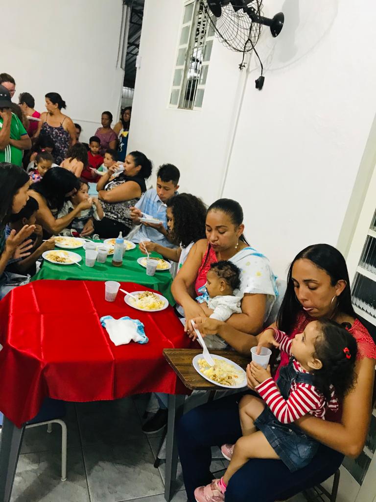 A imagem mostra algumas famílias, sentadas em mesas jantando. Algumas mães dão comida na boca de seus filhos.