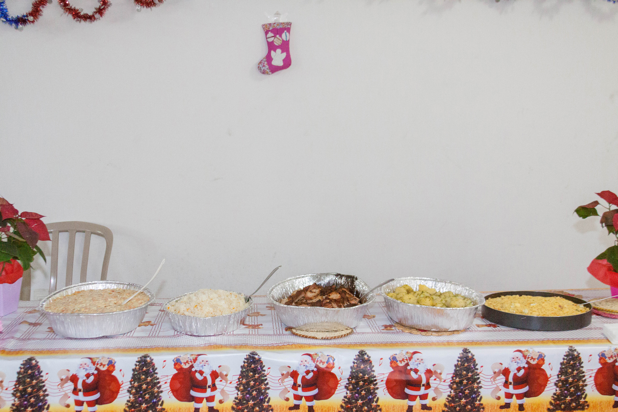 A imagem mostra uma mesa com alguns pratos de natal. Sob a a mesa, há uma toalha com desenhos de papai Noel e árvores natalinas.