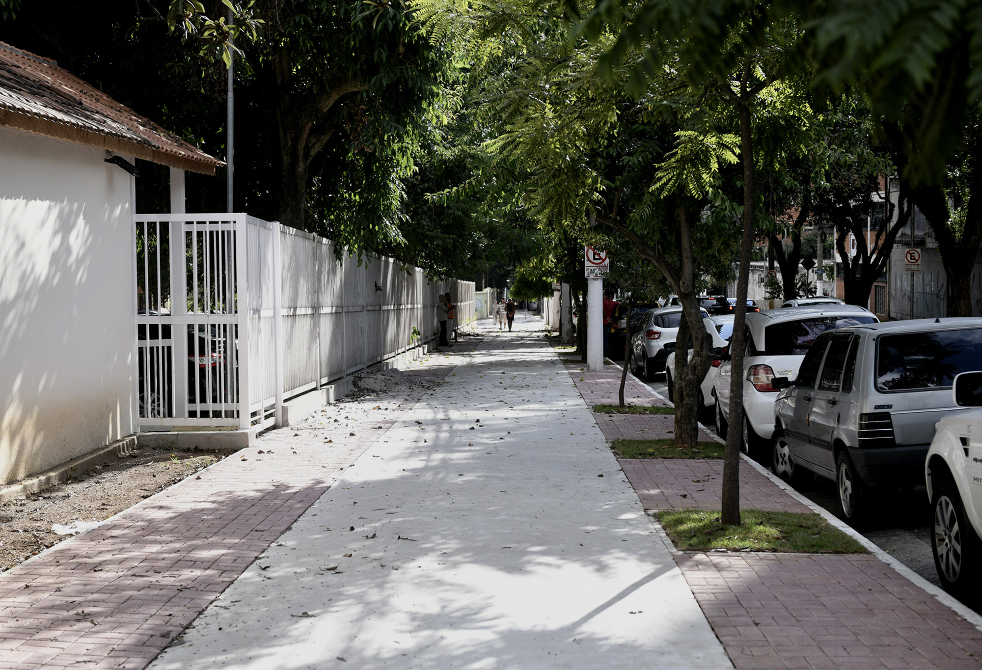 foto de calçada de concreto e piso intertravado nas laterais, à direita duas árvores perfiladas, à esquerda grade de portão branco