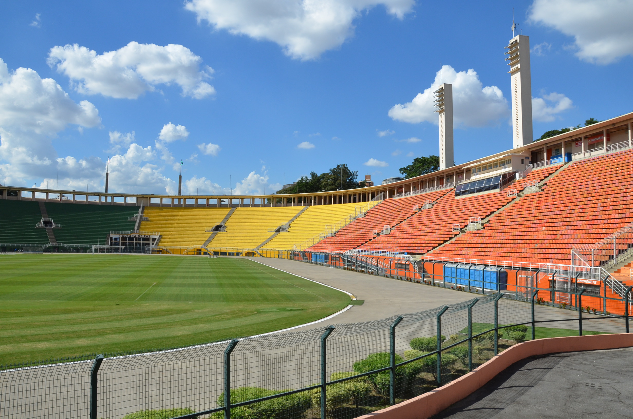 A imagem mostra parte do campo e parte da arquibancada do Estádio do Pacaembu. Estão em destaques os setores laranja e amarelo. O dia está ensolarado, o céu bem azul, com poucas nuvens