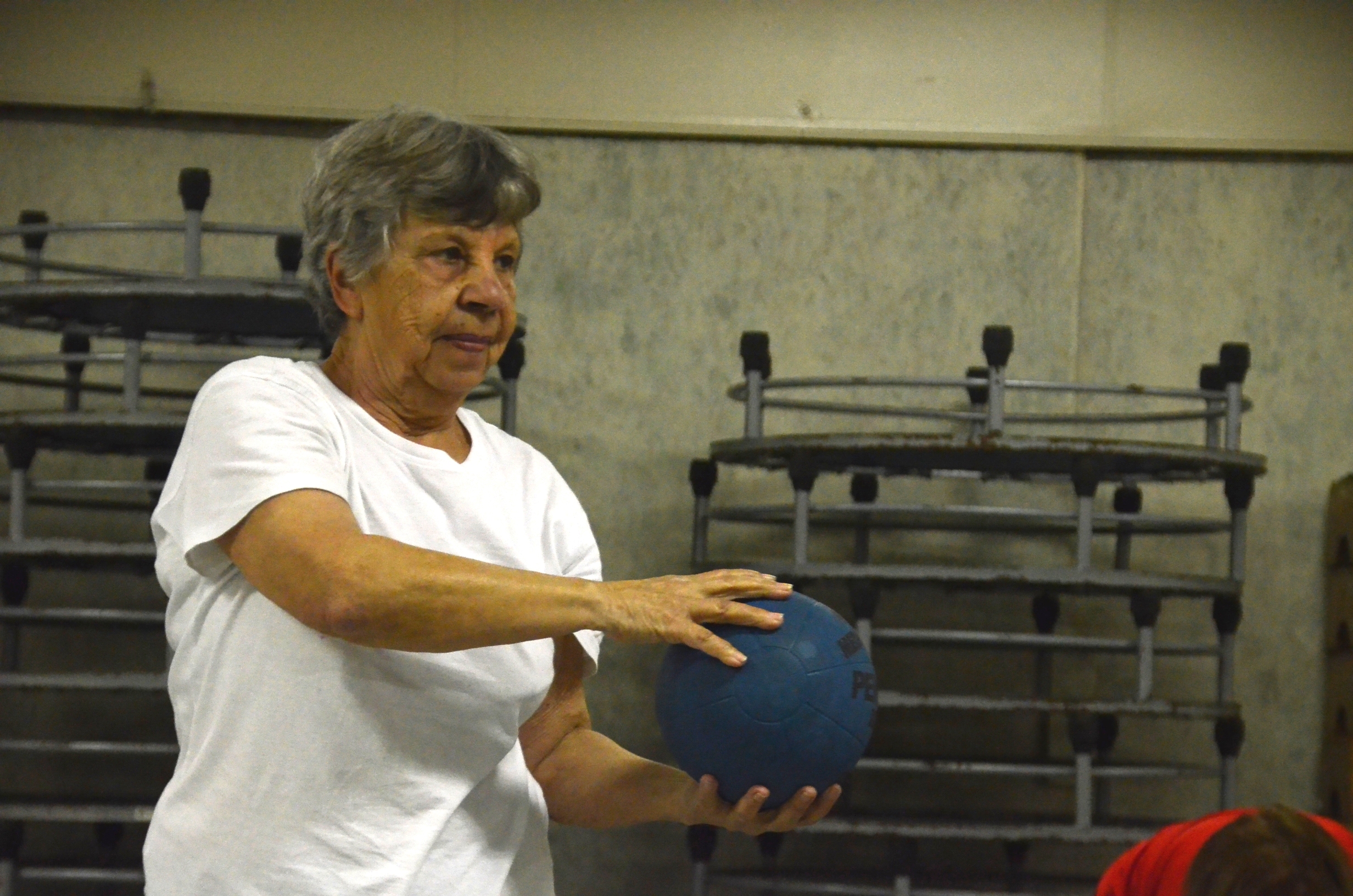 Na imagem, uma idosa está segurando uma bola com as duas mãos, fazendo uma espécie de exercício. Seus braços e a bola azul estão para o lado direito