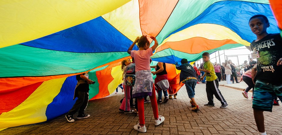 Foto de várias crianças brincando com um tecido colorido em parques municipais.