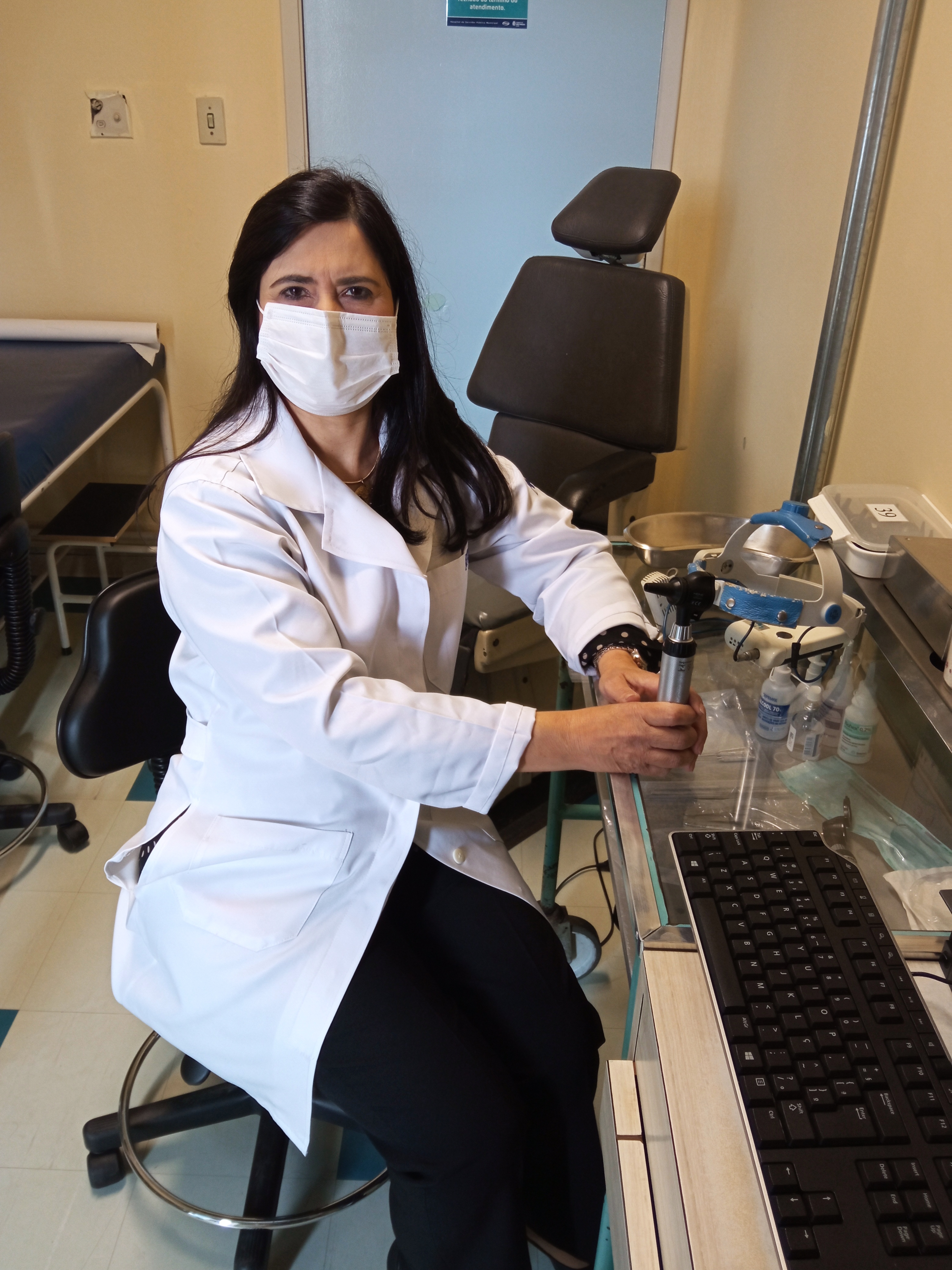 Foto da médica, sentada no consultório do HSPM, com jaleco branco e segurando um equipamento similar a lupa para verificar o interior do ouvido.