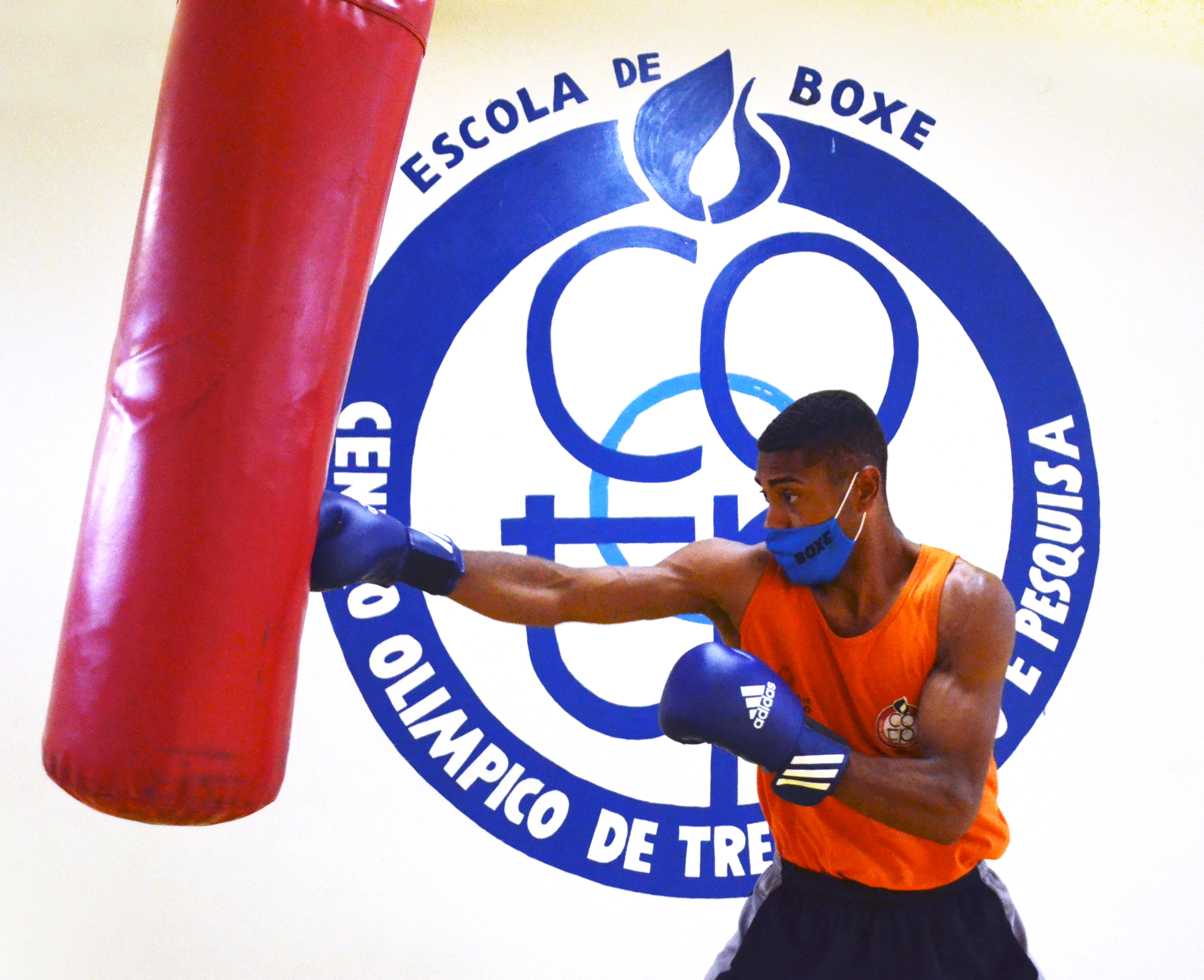 O lutador Luiz Carlos de máscara com uma luva azul, golpeando com as mãos um saco vermelho de boxe.