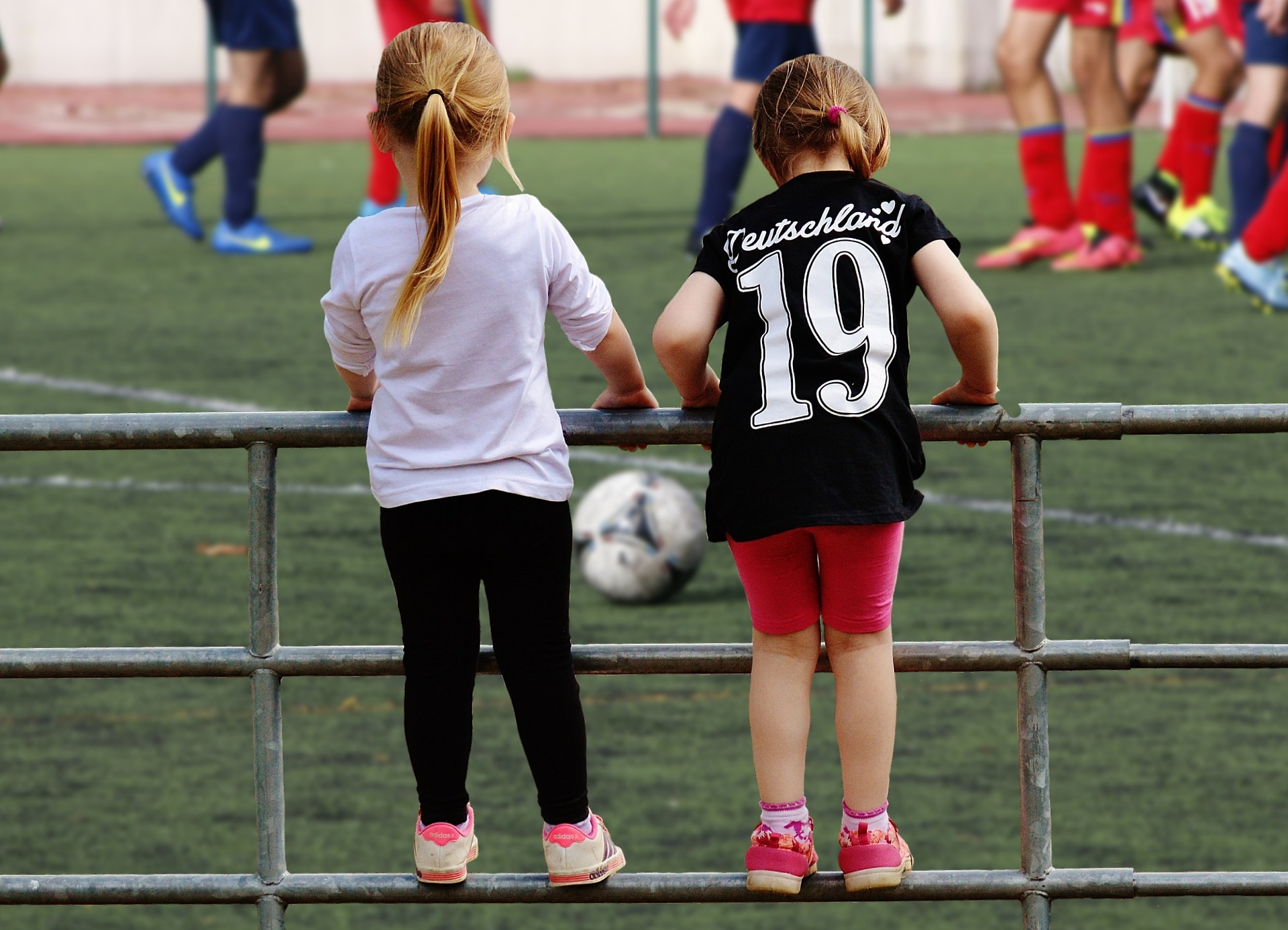 Na imagem, duas meninas aparecem de costas para a câmera, penduradas na grade do campo e olhando em direção à bola e aos jogadores