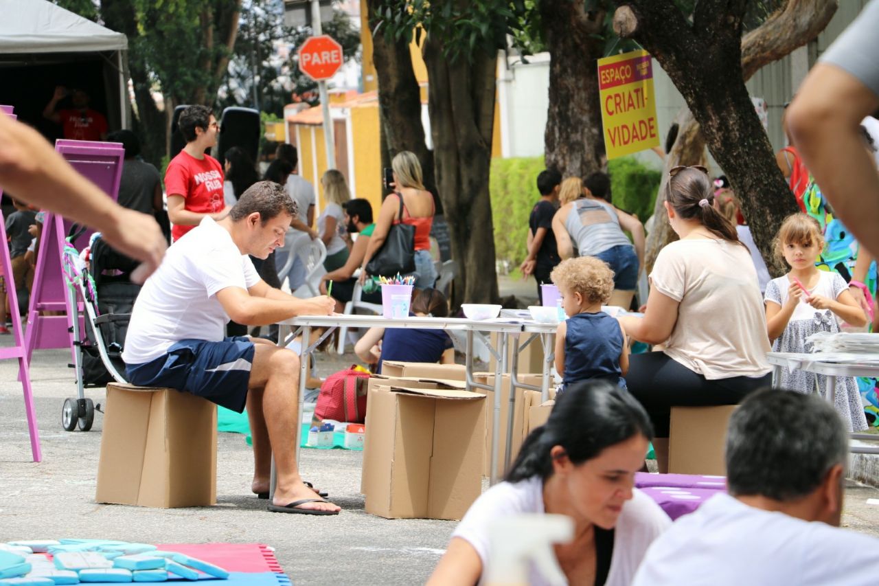 Rua de Lazer em São Paulo. No centro da foto, dois adultos sentados à mesa com uma criança, enquanto pintam algo numa folha no meio do asfalto. Pessoas espalhadas pela rua também compõem a foto.