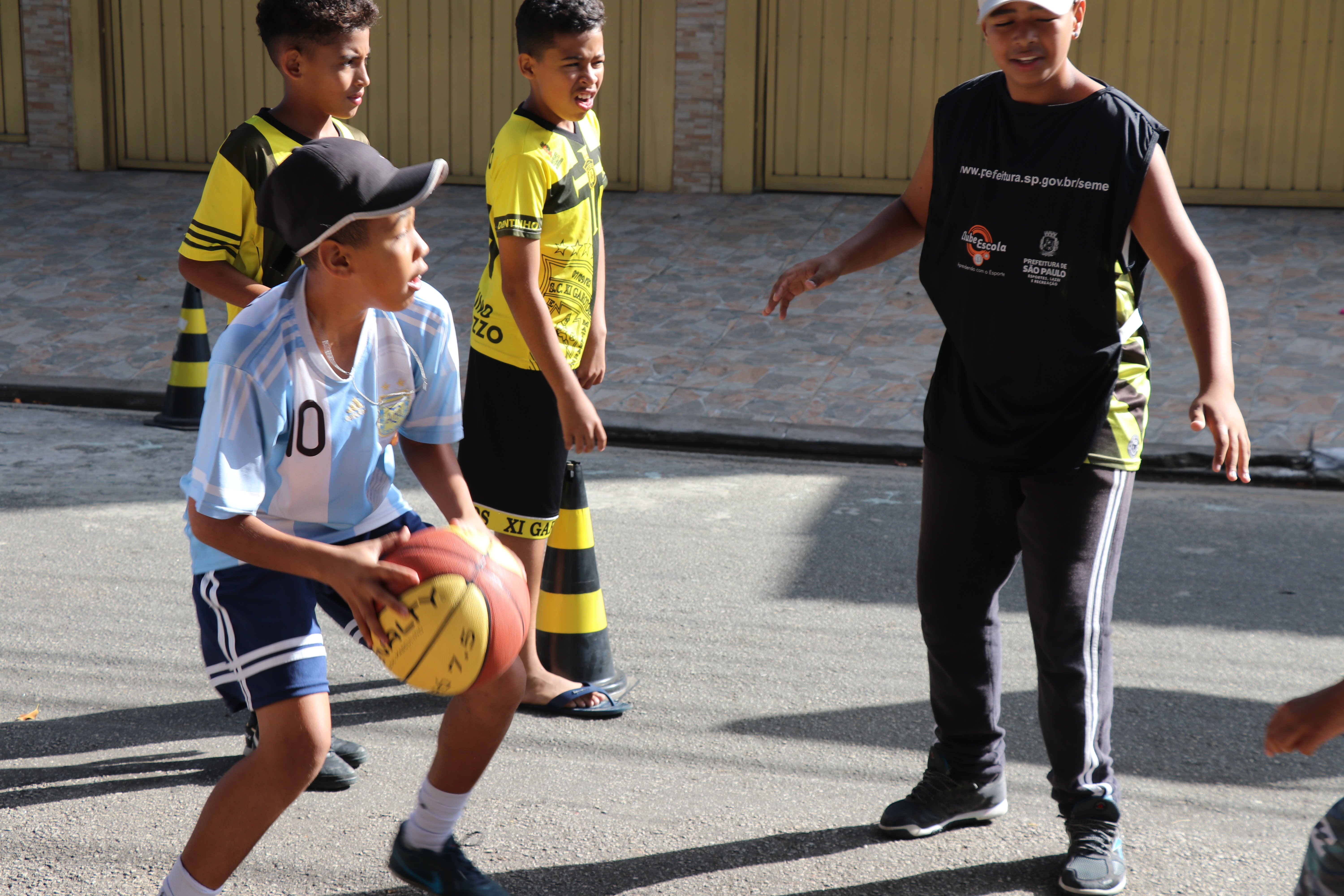 Crianças jogam basquete na rua. Em primeiro plano há um garoto com a bola na mão.