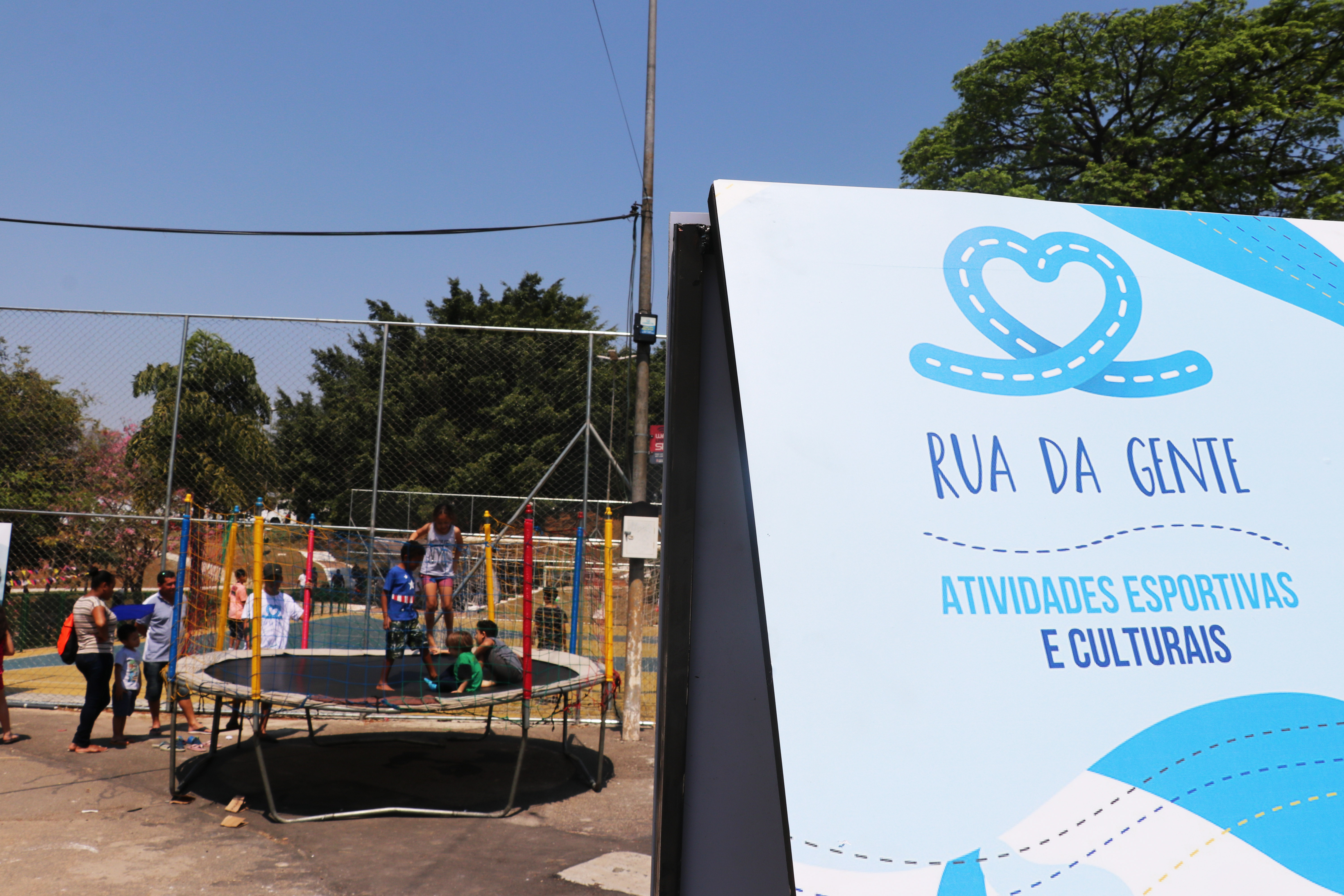 Placa com o logo do Rua da Gente e texto escrito "atividades esportivas e culturais" em primeiro plano. Ao fundo, crianças brincam em um pula-pula.