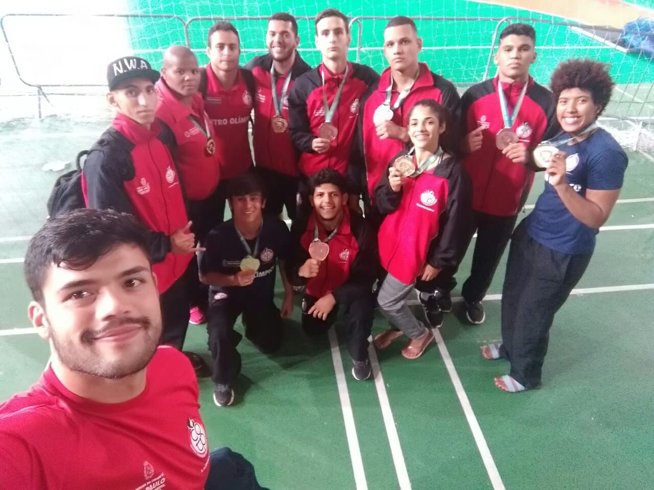 Equipe da Luta Olímpica posa para foto com suas medalhas. Todos estão com o agasalho vermelho do COTP, e o professor está à frente, batendo a selfie.
