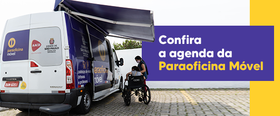 A foto mostra uma van adesivada com o brasão da cidade de São Paulo e da Paraoficina Móvel; ao seu lado, sob um toldo adaptado ao veículo, vê-se uma pessoa em uma cadeira de rodas outra, uma mulher, de pé ao seu lado